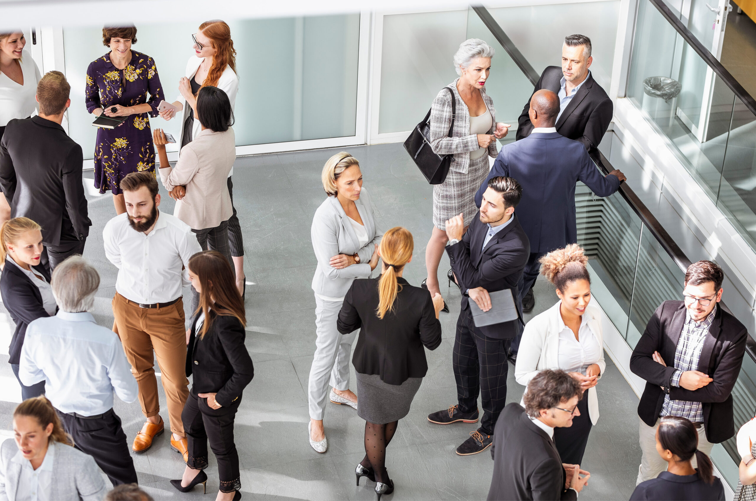 A diverse group of men and women in formal attire engage in animated discussions at a business conference. With notebooks and laptops in hand, they gather in a modern conference hall, fostering collaborative networking and professional growth.